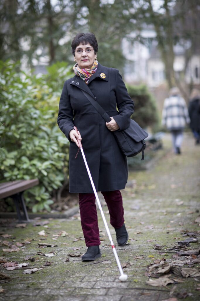 Eine Frau mit Sehbehinderung geht mit einem weißen Langstock über einen Weg im Freien.